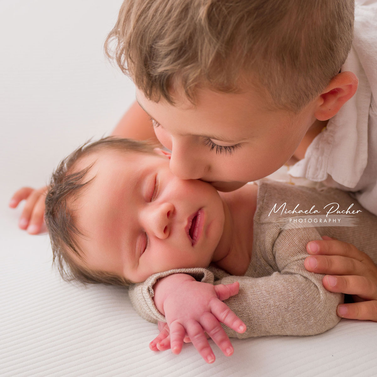 Älteres Geschwisterkind gibt dem schlafenden Neugeborenen einen sanften Kuss auf die Wange, während es beschützend eine Hand auf dessen Rücken legt.